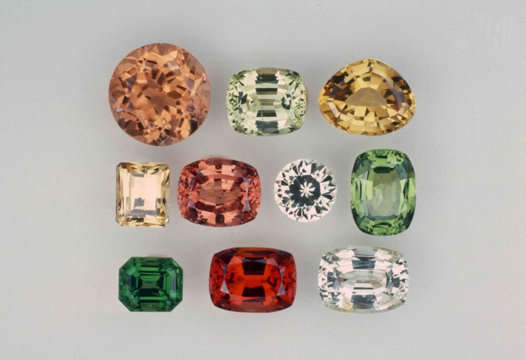 A collection of eleven faceted grossular garnets in various shapes and colors, ranging from cinnamon orange and golden yellow to mint green, emerald green, and colorless.