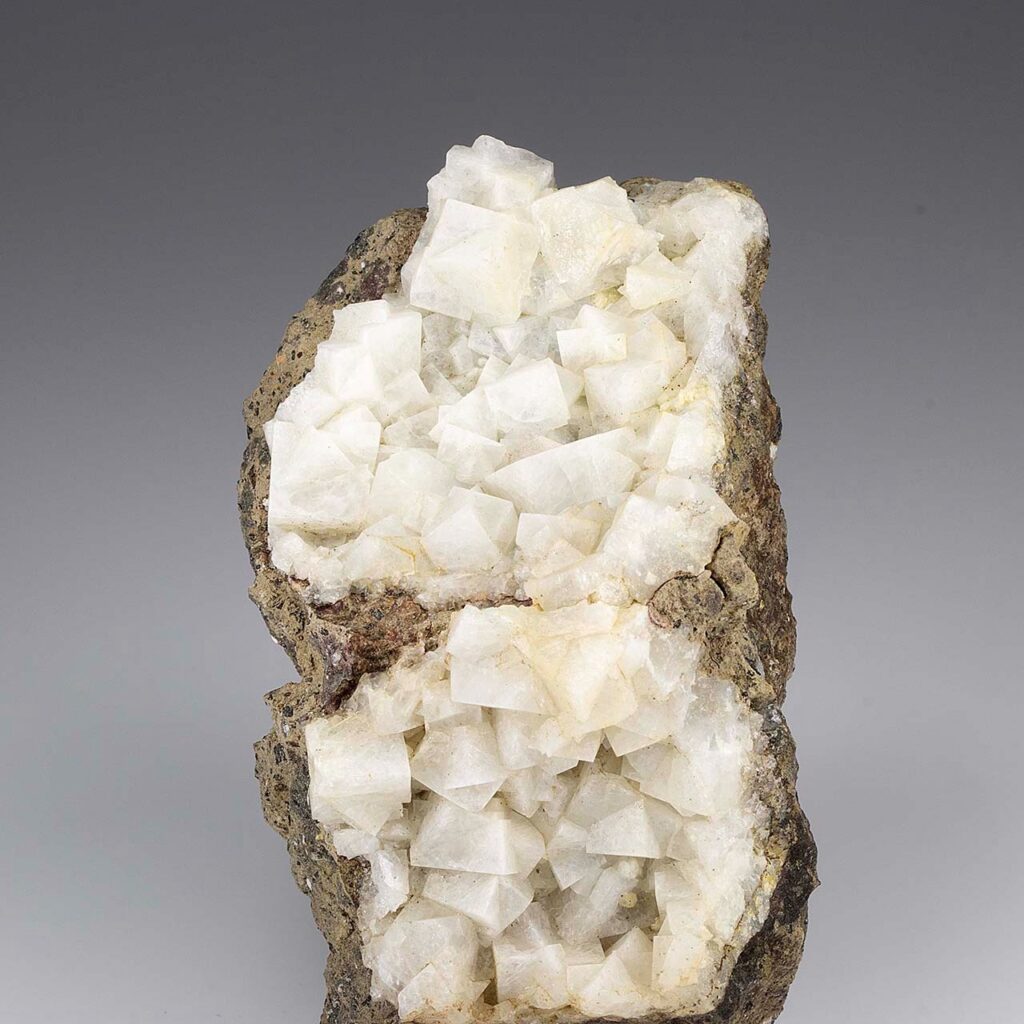 一垂直矿物标本,呈白色半透明的菱沸石晶体,具玻璃光泽,在深色火山岩基质上呈现伪立方菱面体晶形。