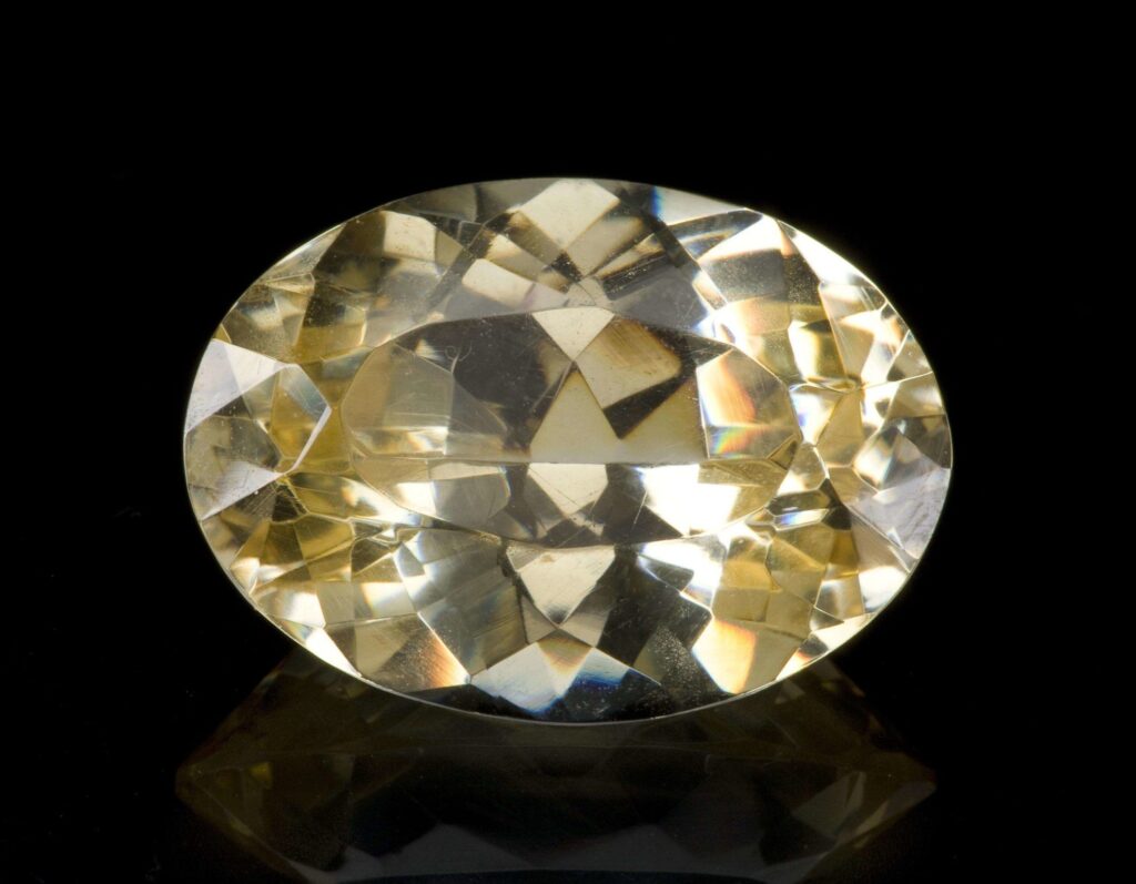 A magnificent 62-carat oval-cut Anglesite gemstone from Morocco. The gem is a bright, pale yellow and displays intense spectral "fire" (dispersion) and brilliance, set against a black reflective background.