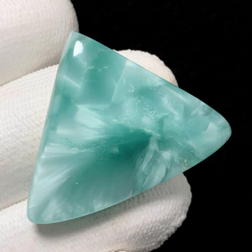 A triangular, cabochon-cut piece of blue-green, translucent Victoria stone, showcasing its characteristic internal white, cloud-like, and feathery patterns.