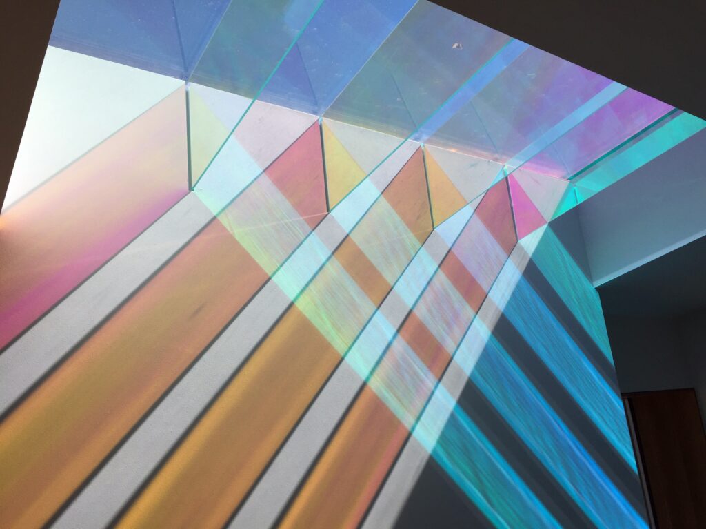 Dichroic glass panels installed in a window, casting vibrant, multi-colored light patterns and prisms of pink, yellow, and blue across an adjacent interior wall.