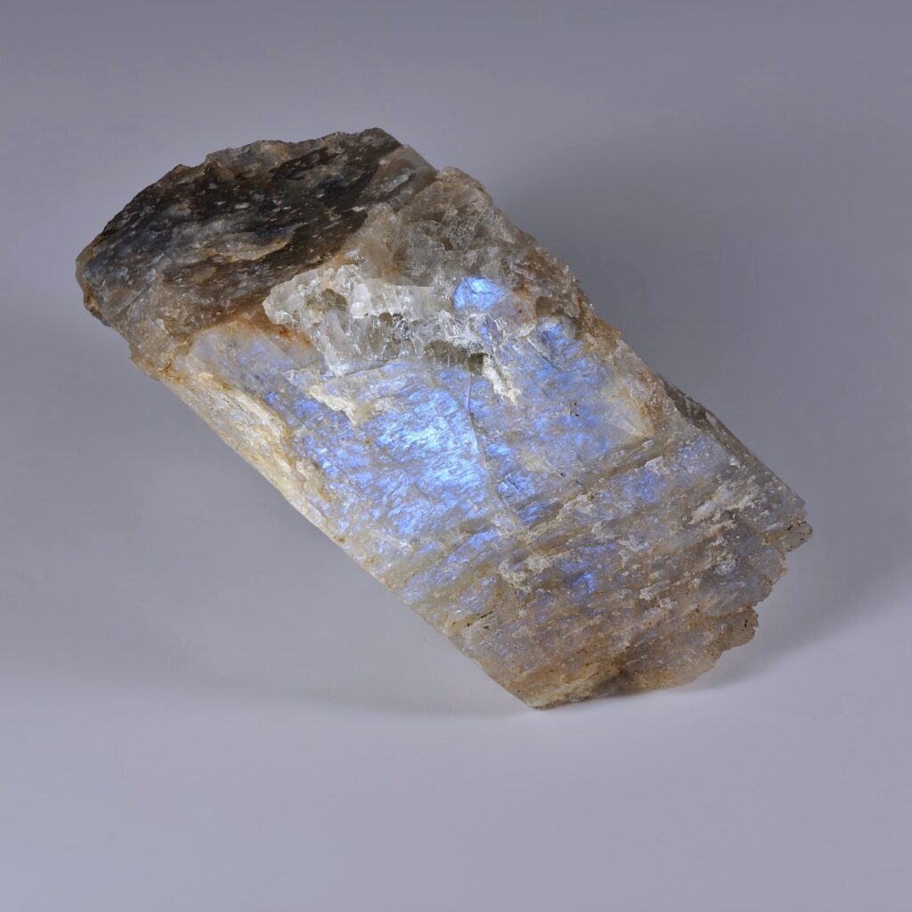 一块钠更长石（奥长石）矿物原石标本，其特点是拥有一个巨大的平整解理面，展现出鲜艳的电光蓝虹彩。该岩石的体色呈半透明白色至棕灰色，具有明显的层状结构，背景为中性灰色。