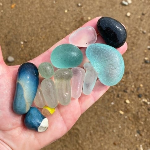 A person's hand holding a collection of various smooth, tumbled pieces of Slag glass in shades of blue, turquoise, pink, and black, with a sandy beach in the background.