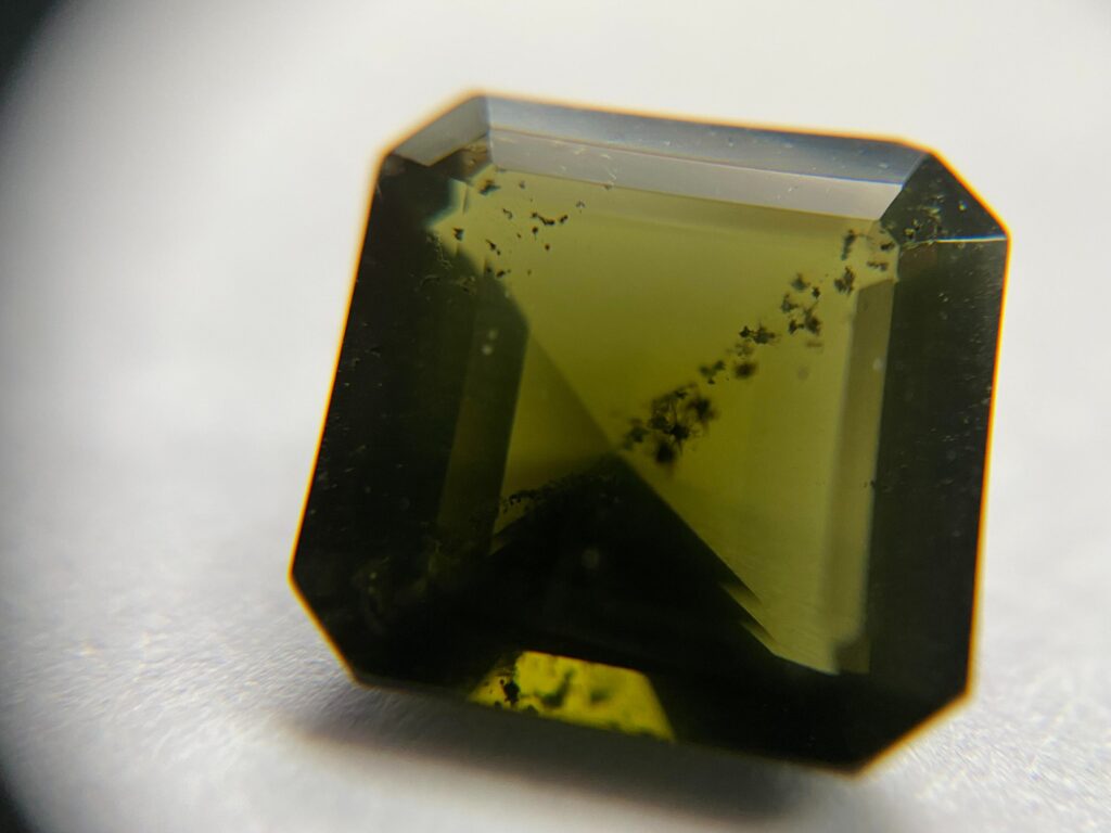 A faceted olive green ekanite gemstone in a square emerald cut, showing characteristic dark internal inclusions.