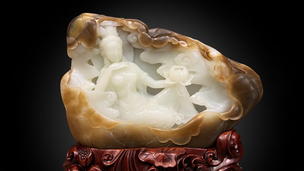 An elaborate Chinese nephrite jade carving set upon a carved wooden stand. The central white jade figures depict Guanyin and an attendant surrounded by clouds and holding a lotus, all enclosed within a natural brown and tan nephrite rind or 'skin' shell against a black gradient background.