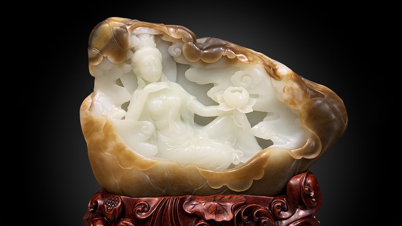 An elaborate Chinese nephrite jade carving set upon a carved wooden stand. The central white jade figures depict Guanyin and an attendant surrounded by clouds and holding a lotus, all enclosed within a natural brown and tan nephrite rind or 'skin' shell against a black gradient background.