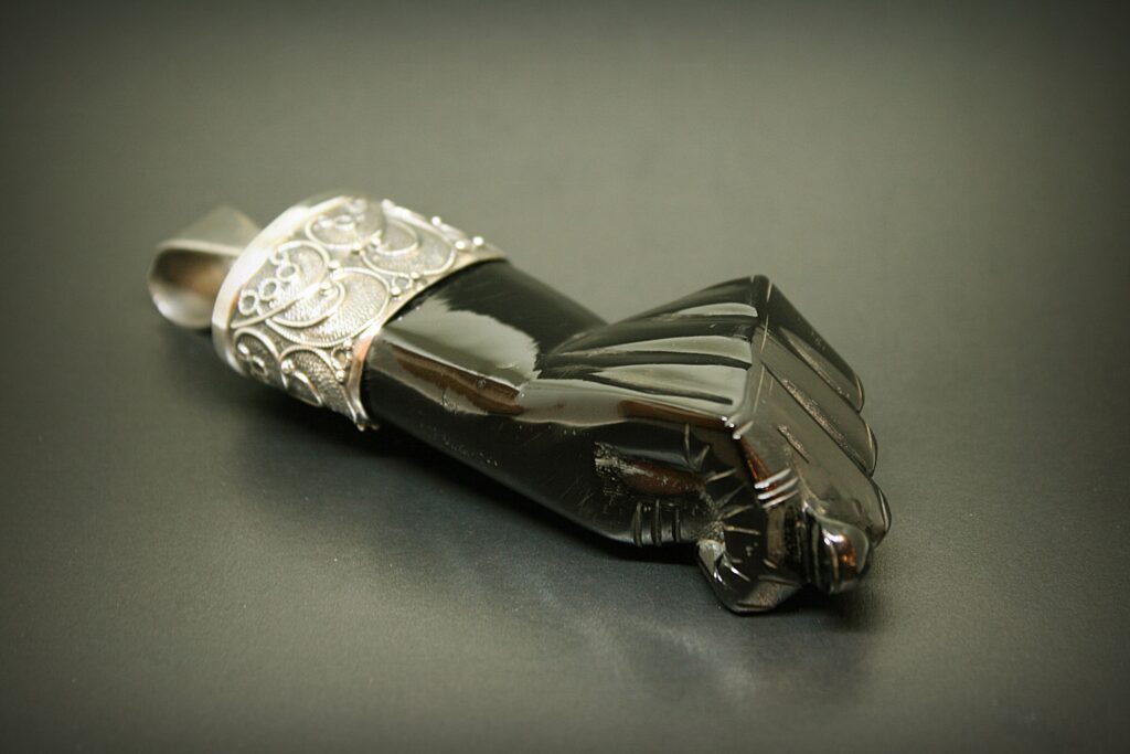 一个传统的 Mano Figa(拳头形状)护身符,由抛光的惠特比煤玉雕刻而成,配有华丽的银色花丝帽,背景为深色。