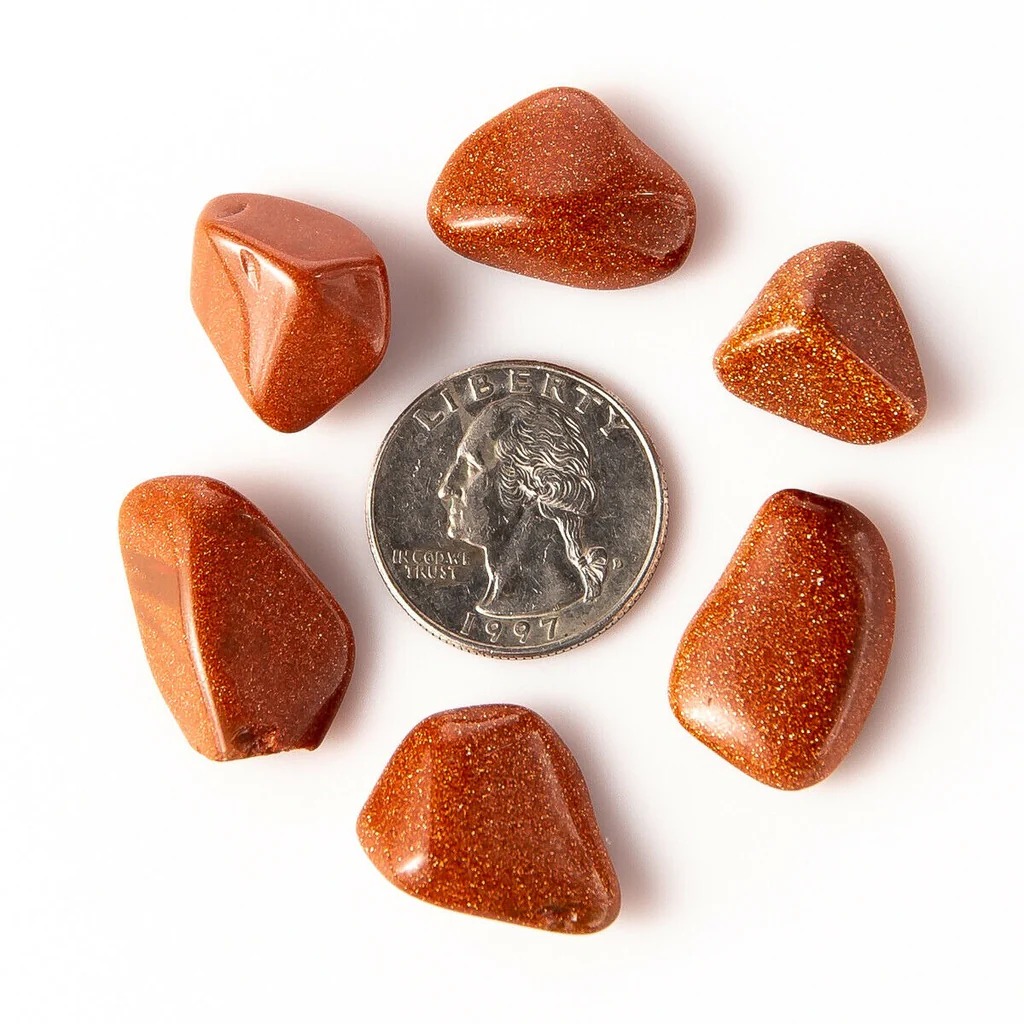 A collection of six tumbled, glittery, reddish-brown Goldstone (Aventurine Glass) stones arranged in a circle around a 1997 United States quarter for scale.