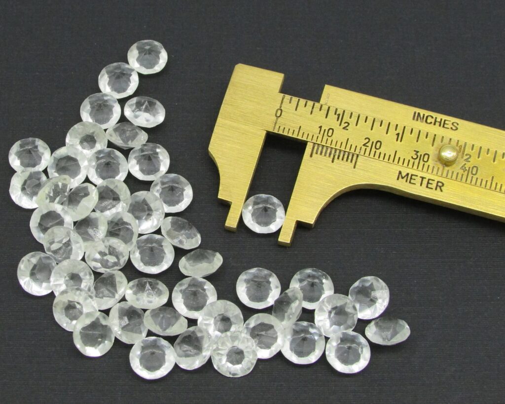A group of small, clear faceted glass stones being measured for size with a golden brass caliper.