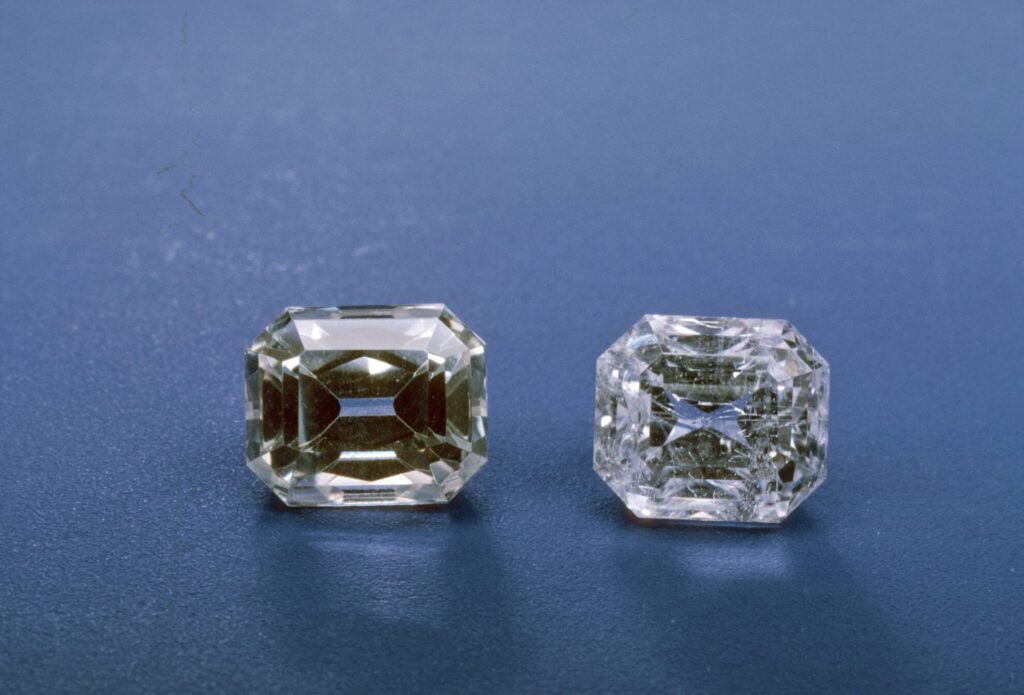 Two emerald-cut Anglesite gemstones resting on a textured blue surface; the left stone is a pale champagne yellow, and the right stone is colorless and transparent.
