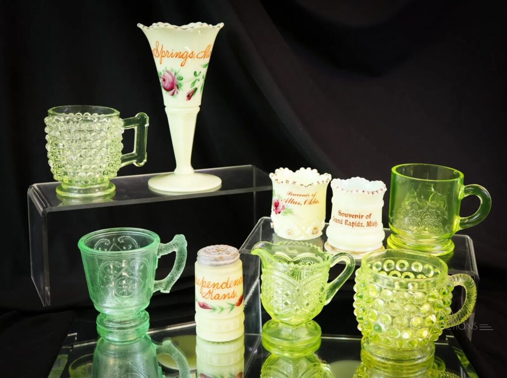 A display of various vintage pressed glass pieces, including mugs, a souvenir vase, and a creamer, all featuring the distinct translucent light green hue characteristic of Uranium or Vaseline glass.