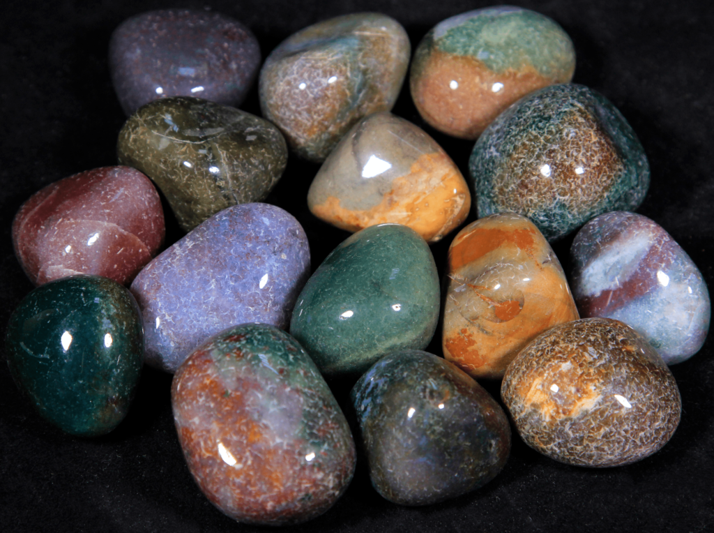 A group of assorted polished Fancy Jasper tumbled stones arranged on a black surface. The collection displays a diverse range of colors, including earthy tones of green, orange, rust, and purple, with unique speckled and swirled patterns on each stone.