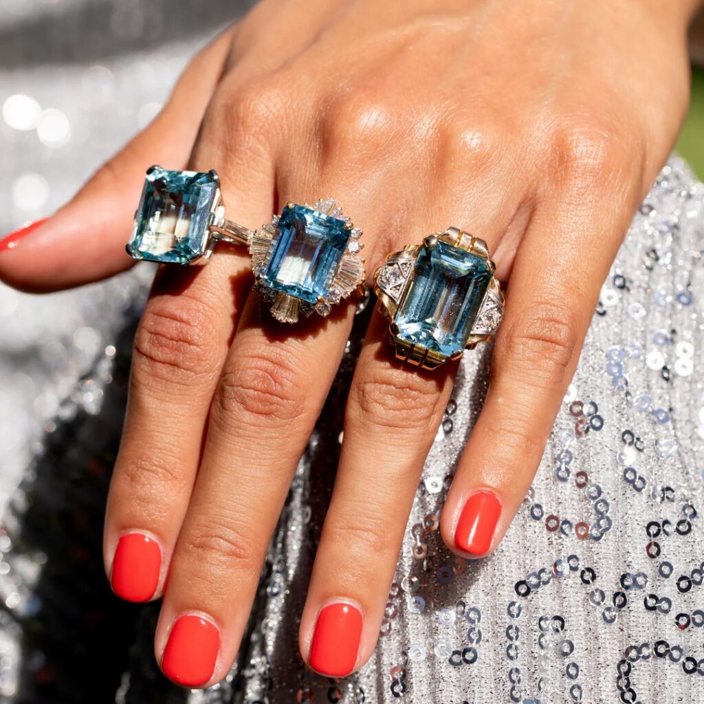 A hand showcasing three different styles of aquamarine cocktail rings with diamond accents