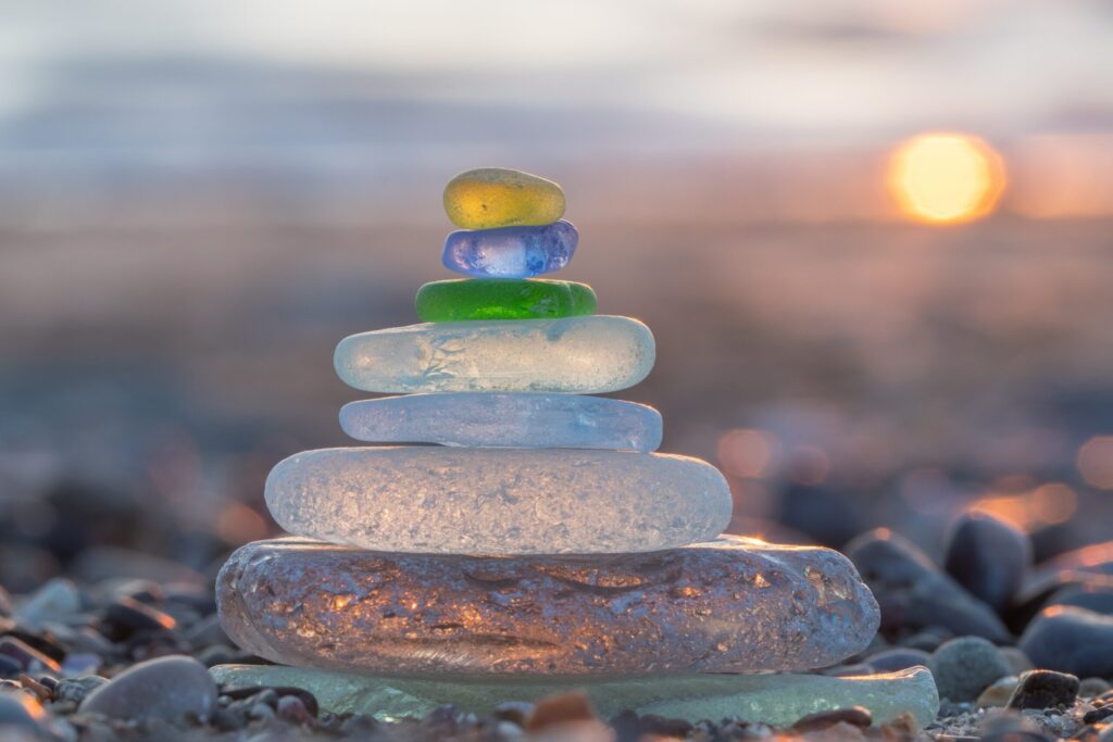 A balanced stack of multi-colored, smooth, and frosted sea glass pieces resting on a sandy beach at sunset, with a blurred, warm background.