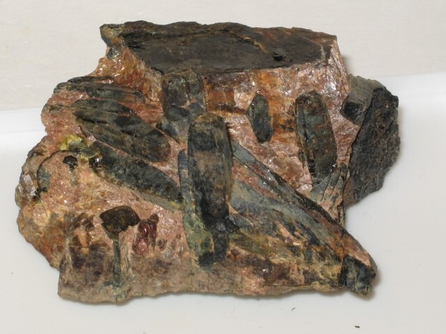 A raw mineral specimen of dark, elongated Fergusonite crystals embedded within a reddish-brown granitic pegmatite matrix.