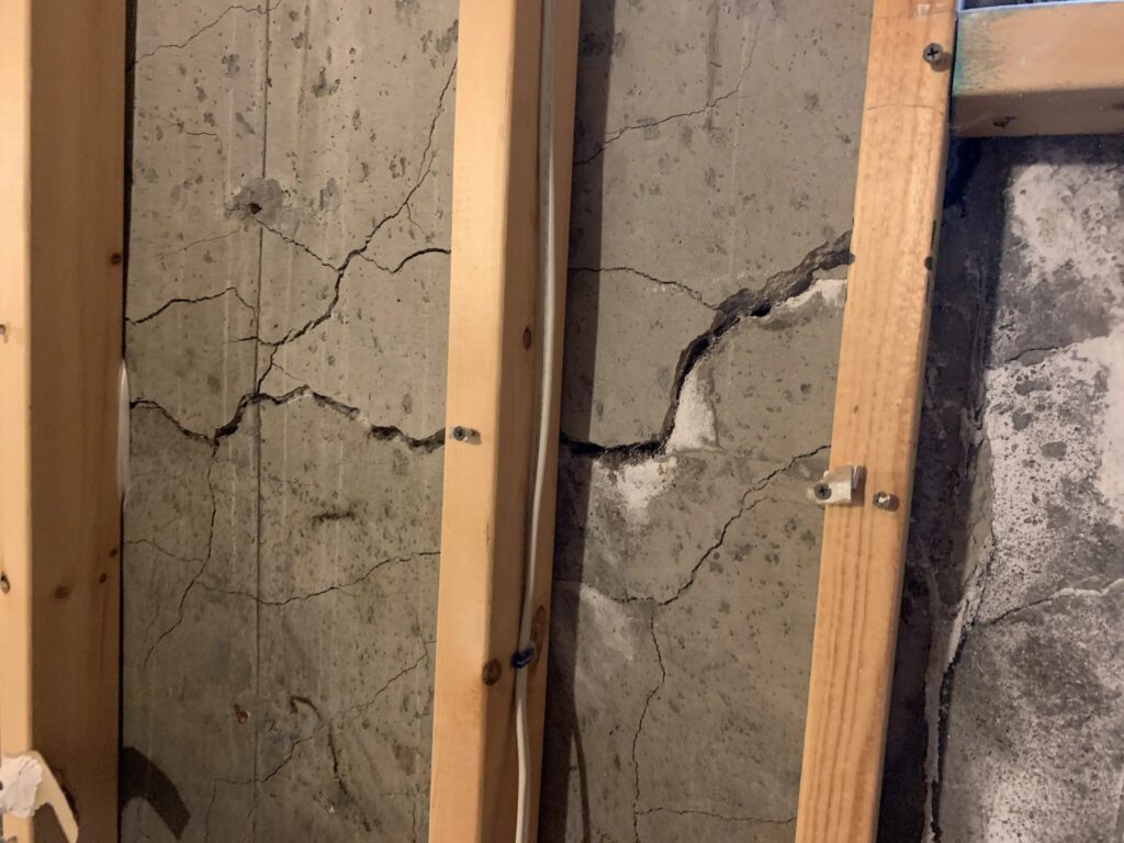 Nahaufnahme einer Betonwand mit schweren netzartigen („Map-Cracking“) und horizontalen Rissen hinter Wandpfosten aus Holz.