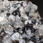 Nahaufnahme einer Mineralprobe mit transparenten bis weißen, glasartigen Anorthit-Kristallen, die mit dunklen, braunschwarzen metallischen Körnern auf einer Gesteinsmatrix vermischt sind.