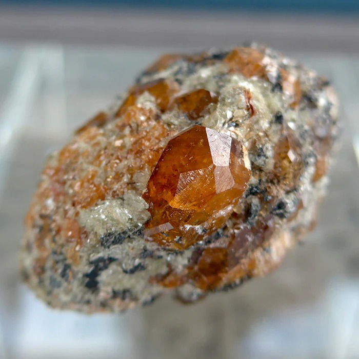 A natural Spessartite Garnet crystal specimen with well-defined dodecahedral faces, embedded in a matrix of granitic pegmatite featuring quartz and mica.