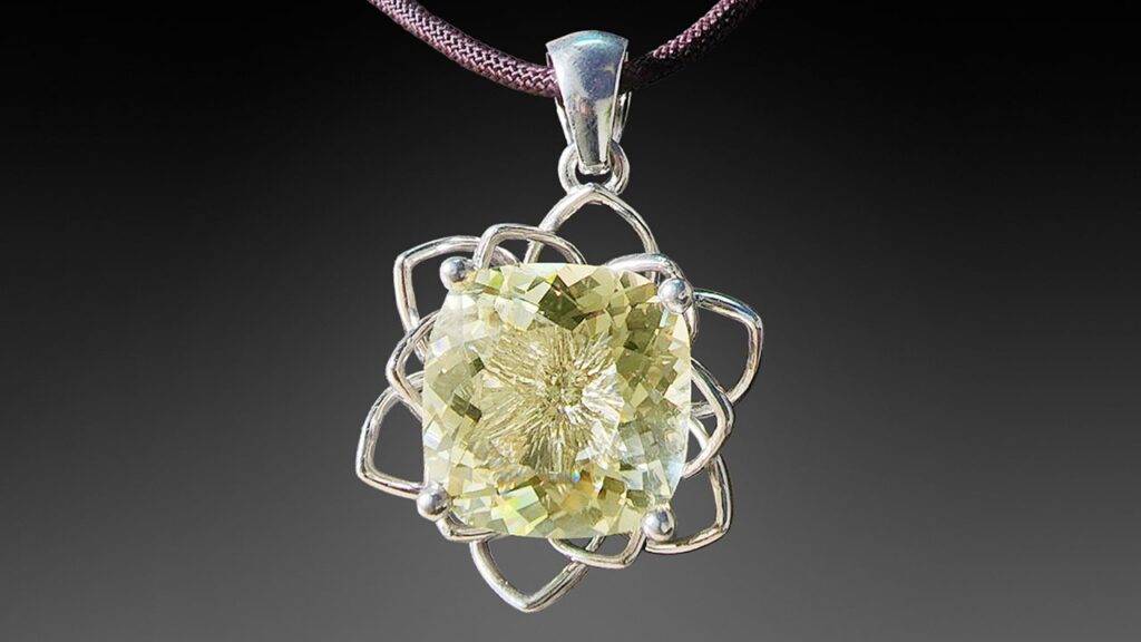 어두운 색상의 끈에 매달린 실버 펜던트로, 꽃이나 기하학적 문양의 정교한 와이어 프레임에 면치기 처리된 연한 황록색 아노르토클레이스 보석이 세팅되어 있습니다.
