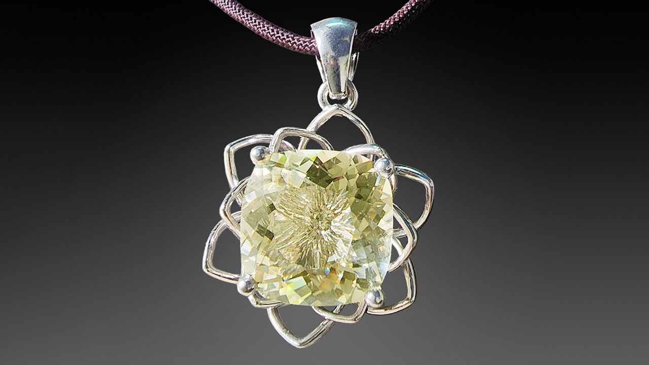 어두운 색상의 끈에 매달린 실버 펜던트로, 꽃이나 기하학적 문양의 정교한 와이어 프레임에 면치기 처리된 연한 황록색 아노르토클레이스 보석이 세팅되어 있습니다.