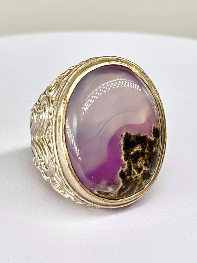 An intricately carved silver ring featuring a rare, oval cabochon-cut purple and pink hodgkinsonite gemstone with distinctive natural dark mineral inclusions.